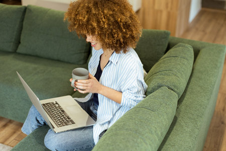 Sitting comfortably on a green sofa, a young woman savors a warm cup of coffee while working on her laptop in a bright, serene living room filled with natural light.の写真素材