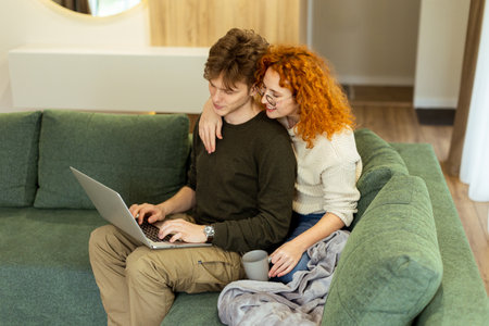 In a stylish living room, a couple shares a warm moment as they sit on a green couch, working on a laptop. The sunlight bathes them, perfect for a relaxed afternoonの写真素材