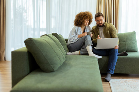 Friends share a joyful moment on a green sofa, discussing ideas while one enjoys a warm drink and the other works on a laptop, surrounded by soft natural light.の写真素材