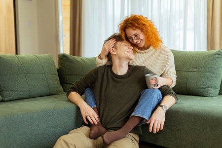 A couple enjoys a relaxing afternoon at home, seated comfortably on a plush sofa, exchanging loving smiles and laughter while sharing a warm beverage togetherの写真素材