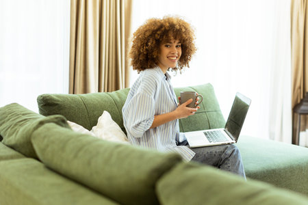 Sitting comfortably on a green sofa, a young woman savors a warm cup of coffee while working on her laptop in a bright, serene living room filled with natural light.の写真素材