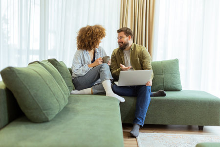 A cheerful couple sits side by side on a plush green couch, engaged in an exciting conversation as they browse through ideas on a laptop under warm sunlight streaming inの写真素材