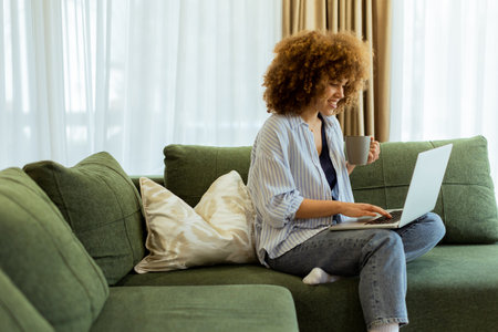 Sitting comfortably on a plush sofa, a young woman savors her warm coffee while working on her laptop, illuminated by soft natural light in a stylish living spaceの写真素材