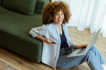 Seated comfortably on the floor, a young woman engages with her laptop while checking her smartphone. The cozy, sunlit living room creates a serene backdrop for her dual focusの写真素材