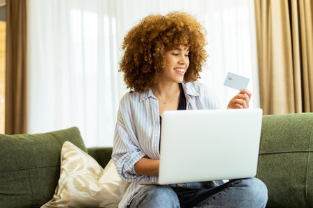A young woman with curly hair happily shops online, holding a credit card, while seated on a comfortable couch in a stylish living room filled with warm sunlight.の写真素材