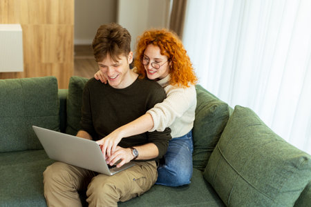 In a stylish living room, a couple shares a warm moment as they sit on a green couch, working on a laptop. The sunlight bathes them, perfect for a relaxed afternoonの写真素材