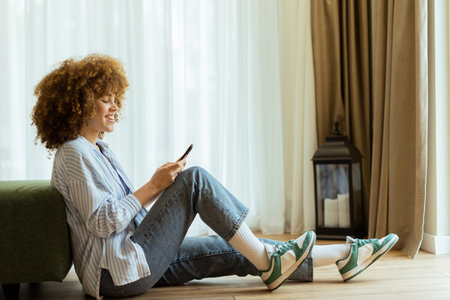 A young woman with curly hair sits on the wooden floor, smiling while looking at her smartphone in a sunlit room. Soft curtains sway gently as she enjoys her peaceful momentの写真素材