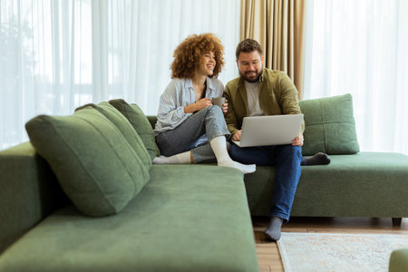 A cheerful couple sits side by side on a plush green couch, engaged in an exciting conversation as they browse through ideas on a laptop under warm sunlight streaming inの写真素材