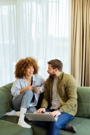 A cheerful couple sits side by side on a plush green couch, engaged in an exciting conversation as they browse through ideas on a laptop under warm sunlight streaming inの写真素材