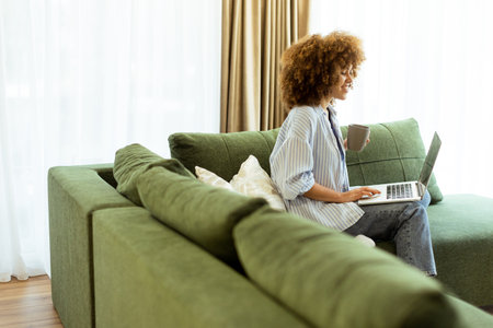 Sitting comfortably on a green sofa, a young woman savors a warm cup of coffee while working on her laptop in a bright, serene living room filled with natural light.の写真素材