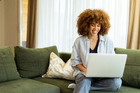 Sitting comfortably on a plush green sofa, a young woman types on her laptop, basking in the warm sunlight streaming through the large windows, radiating focus and tranquility.の写真素材