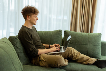 A young man sits comfortably on a green sofa, deeply focused on his laptop. Sunlight pours through large windows, creating a warm atmosphere in the inviting space.の写真素材