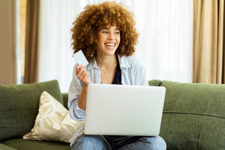A young woman with curly hair happily shops online, holding a credit card, while seated on a comfortable couch in a stylish living room filled with warm sunlight.の写真素材