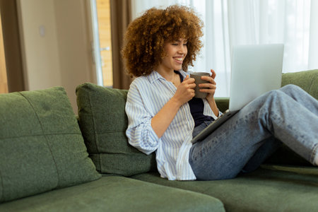 Sitting comfortably on a plush sofa, a young woman savors her warm coffee while working on her laptop, illuminated by soft natural light in a stylish living spaceの写真素材