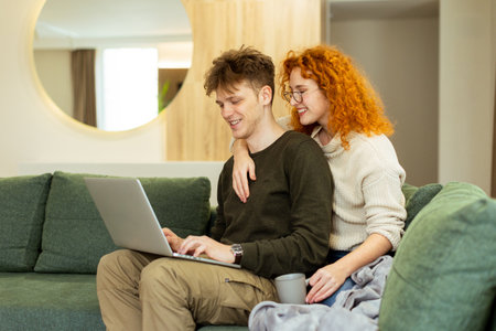 In a stylish living room, a couple shares a warm moment as they sit on a green couch, working on a laptop. The sunlight bathes them, perfect for a relaxed afternoonの写真素材