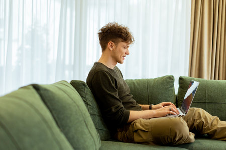 A young man sits comfortably on a green sofa, deeply focused on his laptop. Sunlight pours through large windows, creating a warm atmosphere in the inviting space.の写真素材