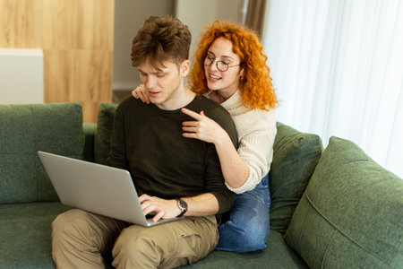 In a stylish living room, a couple shares a warm moment as they sit on a green couch, working on a laptop. The sunlight bathes them, perfect for a relaxed afternoonの写真素材