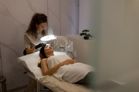 Under soft lighting, a skilled skincare specialist gently performs a treatment on a client lying comfortably on a treatment bed, emphasizing relaxation and rejuvenation.の写真素材