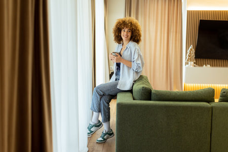 A young individual with curly hair sits on a green sofa, holding a warm cup of coffee, smiling while enjoying the serene ambiance of a stylish living space in soft morning lightの写真素材