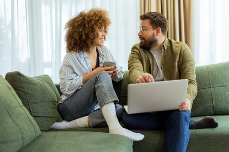 Friends share a joyful moment on a green sofa, discussing ideas while one enjoys a warm drink and the other works on a laptop, surrounded by soft natural light.の写真素材