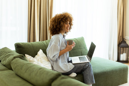 Sitting comfortably on a green sofa, a young woman savors a warm cup of coffee while working on her laptop in a bright, serene living room filled with natural light.の写真素材