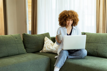 Sitting comfortably on a green sofa, a young woman savors a warm cup of coffee while working on her laptop in a bright, serene living room filled with natural light.の写真素材