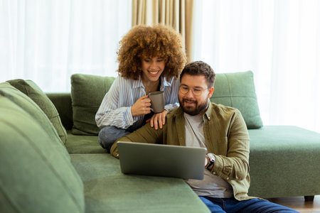 A cheerful couple sits side by side on a plush green couch, engaged in an exciting conversation as they browse through ideas on a laptop under warm sunlight streaming inの写真素材