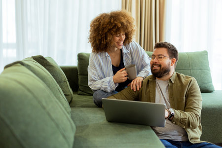 Friends share a joyful moment on a green sofa, discussing ideas while one enjoys a warm drink and the other works on a laptop, surrounded by soft natural light.の写真素材