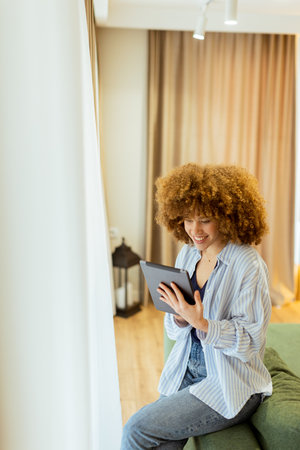 A woman with curly hair enjoys using her tablet, seated comfortably on a couch in a bright, inviting living space, surrounded by soft curtains and warm lightingの写真素材