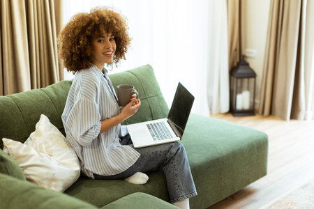 Sitting comfortably on a green sofa, a young woman savors a warm cup of coffee while working on her laptop in a bright, serene living room filled with natural light.の写真素材