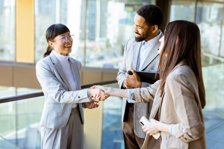Three professionals in stylish business attire share a friendly handshake while discussing ideas in a bright, contemporary office environment with city views.の写真素材