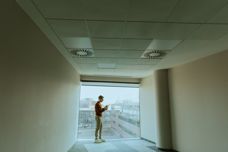 On the upper floor of a modern building, a man with glasses stands alone, focused on his mobile...