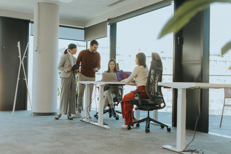 Four professionals engage in a dynamic discussion around laptops in a modern office setting filled with natural light. Ideas flow as they collaborate on a project.の写真素材