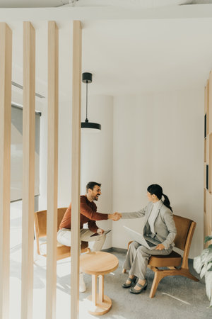 Two individuals in contemporary attire converse intently in a bright lounge featuring wooden accents and greenery. The sunlight casts soft shadows, adding warmth to their discussion.の写真素材