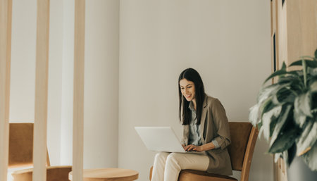 In a bright, minimalist setting, a woman engages joyfully with her laptop, exuding creativity and focus. Soft natural light enhances the simple elegance of her surroundings.の写真素材