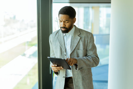 A well-dressed man reflects on his ideas while gazing through a large window in a contemporary office. Sunlight illuminates the space, creating a serene atmosphere for inspiration.の写真素材
