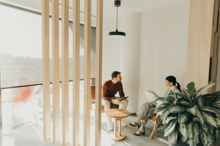 Two individuals in contemporary attire converse intently in a bright lounge featuring wooden accents and greenery. The sunlight casts soft shadows, adding warmth to their discussion.の写真素材