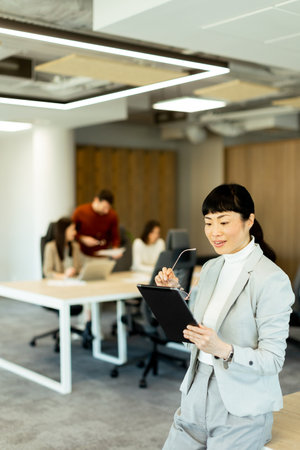 In a modern office, a focused businesswoman in a stylish suit uses her tablet while team members collaborate in the background, fostering a productive atmosphere.の写真素材