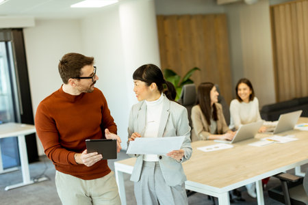 A lively exchange unfolds in a bright office as two professionals collaborate. In the background, coworkers focus on their laptops, enhancing the atmosphere of teamwork and innovation.の写真素材