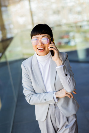 A professional Asian woman in a stylish suit chats joyfully on her phone within a sleek office building with large windows. Her bright smile reflects her positive energy and success.の写真素材