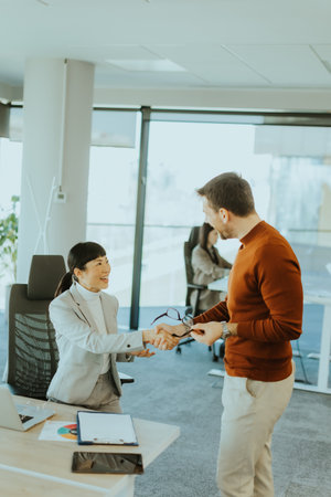 Bright office environment sees a joyful handshake between a male and female professional. They exchange smiles, indicating a fruitful collaboration after discussion at a sleek desk.の写真素材