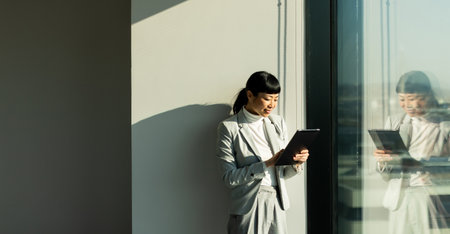 In a sleek office space, Japanese woman in professional attire stands by a large window, interacting with her tablet while basking in the warm glow of sunlight, reflecting her focused demeanor.の写真素材