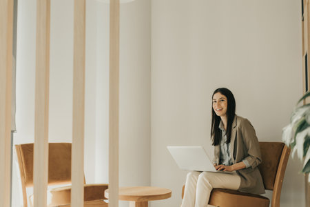 A young woman sits on a stylish chair, engaged in her work on a laptop, surrounded by a serene and minimalistic office design, revealing a calm afternoon atmosphere.の写真素材