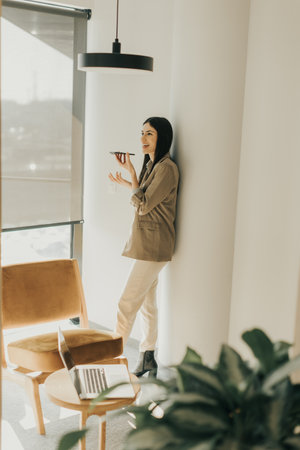 A young woman leans against a wall, smiling as she talks on the phone. Sunlight pours through the window, highlighting her natural beauty and the tranquil atmosphere of the space.の写真素材