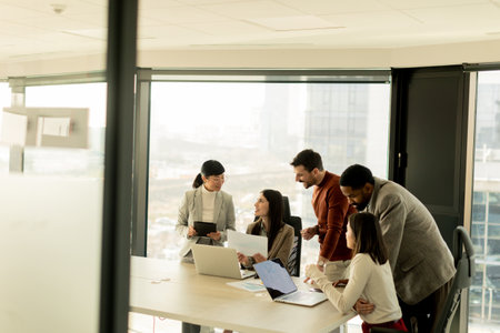 A diverse group of professionals engages in a dynamic brainstorming session around a conference table, sharing ideas while interacting with laptops and notes in a bright office space.の写真素材