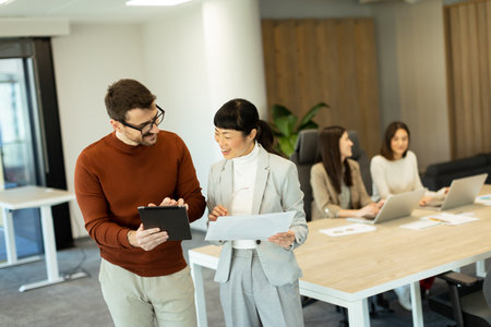 A lively exchange unfolds in a bright office as two professionals collaborate. In the background, coworkers focus on their laptops, enhancing the atmosphere of teamwork and innovation.の写真素材