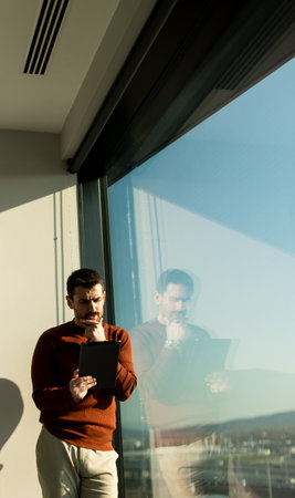 A man stands thoughtfully beside a large window, holding a tablet as he gazes out at the vibrant cityscape. The natural light highlights his focused expression in the modern setting.の写真素材