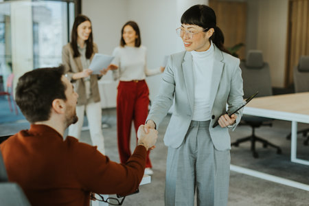 A confident woman shakes hands with a man in a stylish office while colleagues observe, fostering...
