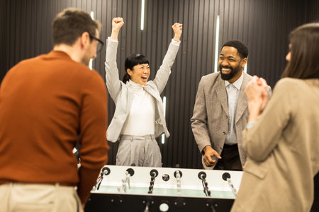 Laughter fills the air as friends cheer during an exciting foosball match in a modern indoor space. One player raises their arms in triumph, showcasing pure joy and camaraderie.の写真素材