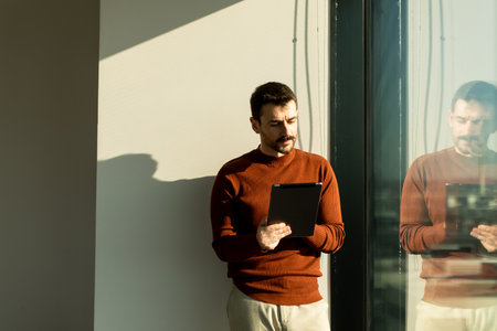 A man engages with a tablet while smiling, standing by a large window in a contemporary office. The...